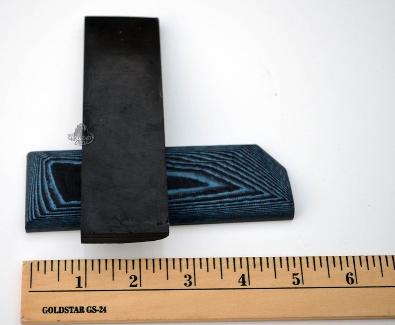 May include: Two black and blue wood blanks for knife making. The blanks are approximately 2 inches long and 1 inch wide. The ruler in the image is marked in inches.