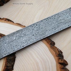May include: A close-up of a long, rectangular piece of metal with a swirling, patterned surface. The metal has a silver colour and a smooth, polished finish. The metal is lying on a wooden surface.