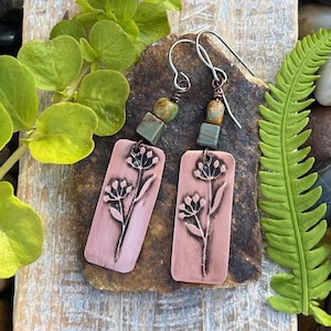 May include: A pair of handcrafted earrings featuring rectangular copper pendants with a floral design. The earrings have silver-colored ear wires and are accented with green and brown beads. The earrings are displayed on a stone surface with greenery.