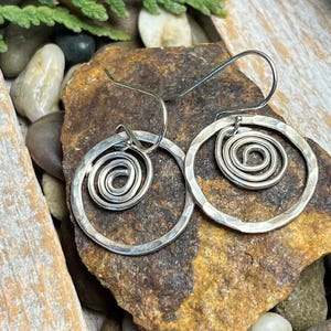 May include: A pair of silver-toned spiral hoop earrings. Each earring features a hammered texture on the outer ring and a coiled spiral design in the center. The earrings are displayed on a textured brown rock surface.