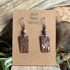 May include: Copper earrings with a rectangular pendant featuring a rose design. The earrings have a beaded accent with brown and green beads. The text "Grape Twine Creations" is printed on the card.