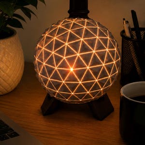May include: A spherical lamp featuring a geometric pattern of raised triangles on its surface. The structure is illuminated from within, casting a warm glow through the grid lines. It is supported by a dark, three-legged base and has a cylindrical top.