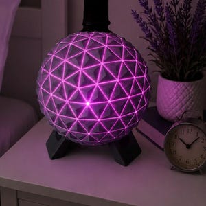 May include: A spherical lamp featuring a geometric geodesic dome pattern with a vibrant pink light emanating from the crevices. The lamp is supported by a black tripod base and has a black cylindrical top component, creating a modern lighting fixture.