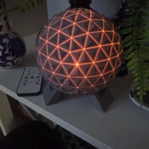 May include: A spherical lamp featuring a geometric geodesic pattern of interconnected triangles. The surface is grey with warm orange light glowing from the seams between the triangular panels. The lamp is supported by a grey three-legged base.