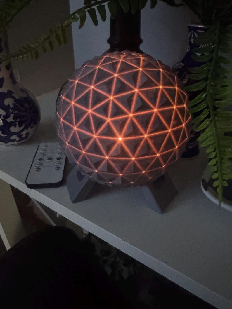 May include: A spherical lamp featuring a geometric geodesic pattern of interconnected triangles. The surface is grey with warm orange light glowing from the seams between the triangular panels. The lamp is supported by a grey three-legged base.