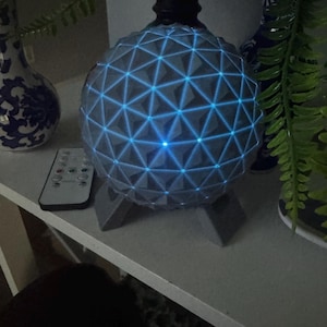 May include: A spherical lamp featuring a gray geodesic pattern with glowing blue LED lights integrated into the structure. The lamp is supported by a gray tripod base and is accompanied by a small rectangular remote control with multiple buttons.