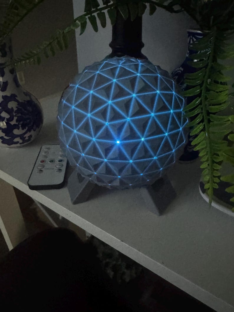 May include: A spherical lamp featuring a gray geodesic pattern with glowing blue LED lights integrated into the structure. The lamp is supported by a gray tripod base and is accompanied by a small rectangular remote control with multiple buttons.