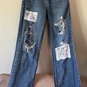 May include: Blue denim jeans with white lace patches and trim. The jeans have distressed details and are a relaxed fit.