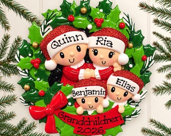 Könnte beinhalten: Ein runder Weihnachtsschmuck mit vier Zeichentrickfiguren, die rote Glitzer-Weihnachtsmützen mit den Namen Quinn, Ria, Benjamin und Elias tragen. Die Figuren sind von grünen Stechpalmenblättern, roten Beeren und einem roten Banner mit der Aufschrift 2026 umgeben.
