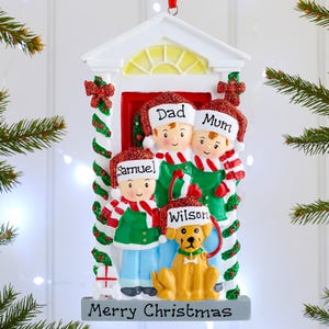 May include: A hanging Christmas ornament shaped like a doorway featuring a family of three and a yellow dog. The figures wear red and white hats with the names Dad, Mum, Samuel, and Wilson. The base is grey with the text Merry Christmas.