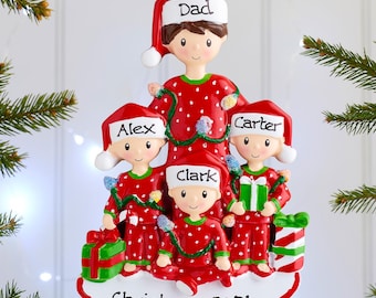 May include: A Christmas tree ornament featuring four figures in red pajamas with white polka dots and Santa hats. The figures are labeled Dad, Alex, Carter, and Clark. The base is white with the text Christmas 2021 in black script lettering.