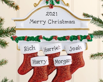 Puede incluir: Un adorno navideño de repisa de chimenea en blanco y dorado con cinta roja, guirnalda verde con bayas rojas y cinco calcetines rojos brillantes. La repisa incluye la inscripción 2021, Merry Christmas y los nombres Rich, Harriet, Josh, Harry y Morgan.
