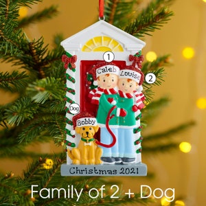 K&ouml;nnte beinhalten: Ein Weihnachtsbaumschmuck in Form eines Hauseingangs mit zwei Figuren in roten M&uuml;tzen namens Caleb und Louise sowie einem gelben Hund namens Bobby. Der Schmuck zeigt eine rote T&uuml;r, gr&uuml;ne Girlanden und den Schriftzug Christmas 2021.