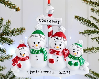 Könnte beinhalten: Ein Weihnachtsschmuck mit vier weißen Schneemännern auf einem verschneiten Sockel mit der Aufschrift Christmas 2021. Die Schneemänner tragen rot-grüne Mützen und Schals mit den Namen Mia, Johnathon, Debbie und Aiden. Darüber befindet sich ein North Pole Schild.