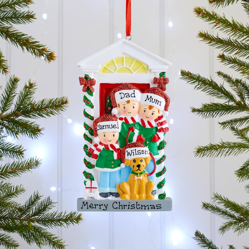 May include: A hanging Christmas ornament shaped like a doorway featuring a family of three and a yellow dog. The figures wear red and white hats with the names Dad, Mum, Samuel, and Wilson. The base is grey with the text Merry Christmas.