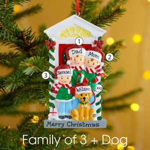 K&ouml;nnte beinhalten: Ein h&auml;ngender Weihnachtsbaumschmuck in Form eines Hauseingangs mit einer dreik&ouml;pfigen Familie und einem Hund. Die Figuren tragen Weihnachtsm&uuml;tzen mit den Namen Dad, Mum, Samuel und Wilson. Der graue Sockel tr&auml;gt die Aufschrift Merry Christmas.