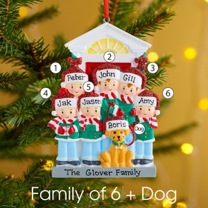 K&ouml;nnte beinhalten: Ein personalisierter Weihnachtsbaumschmuck mit einer sechsk&ouml;pfigen Familie und einem Hund vor einer Hausfassade. Jede Figur tr&auml;gt eine rote Glitzerm&uuml;tze mit einem darauf geschriebenen Namen, darunter Peter, John, Gill, Jak, Jason, Amy und Boris.