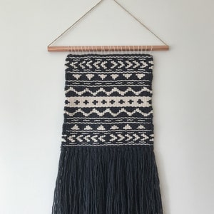 May include: Black and white geometric wall hanging with a fringe border. The hanging is woven with a copper rod at the top.
