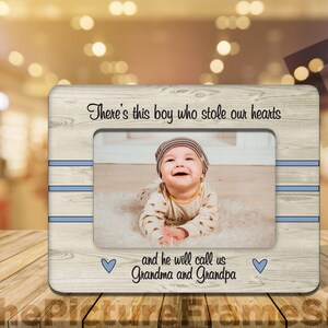 May include: A light wood picture frame with a white and blue striped border. The frame has a photo of a baby boy in it. The text "There's this boy who stole our hearts and he will call us Grandma and Grandpa" is printed on the frame.