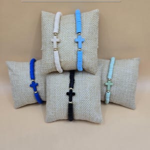 Op de afbeelding: Vijf armbanden met platte ronde kralen en een centraal kruisbedeltje, gepresenteerd op kleine jute kussentjes. De armbanden zijn wit, lichtblauw, donkerblauw, zwart en mintgroen en bevatten goudkleurige tussenkralen. Tekst: Cross Bracelets.