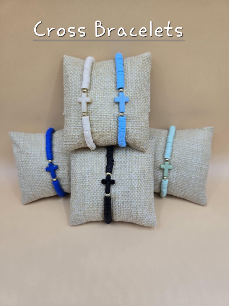 Op de afbeelding: Vijf armbanden met platte ronde kralen en een centraal kruisbedeltje, gepresenteerd op kleine jute kussentjes. De armbanden zijn wit, lichtblauw, donkerblauw, zwart en mintgroen en bevatten goudkleurige tussenkralen. Tekst: Cross Bracelets.