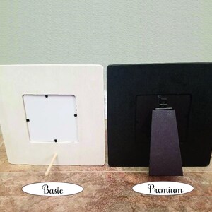 May include: Two square picture frames, one cream-colored and one black, are displayed side-by-side. The cream frame has a simple wooden stand, while the black frame has a more elaborate stand. The word "Basic" is written below the cream frame, and "Premium" below the black frame.