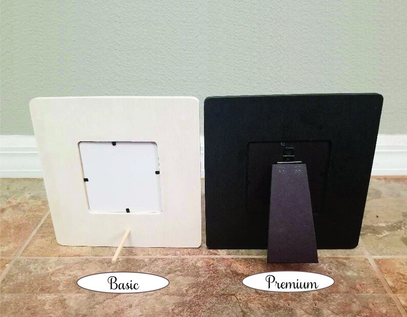 May include: Two square picture frames, one cream-colored and one black, are displayed side-by-side. The cream frame has a simple wooden stand, while the black frame has a more elaborate stand. The word "Basic" is written below the cream frame, and "Premium" below the black frame.