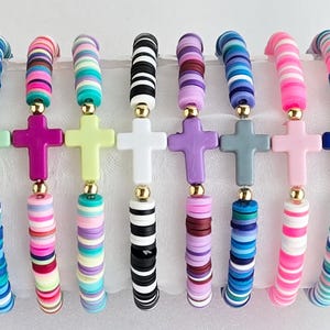 May include: A collection of colorful beaded bracelets. Each bracelet features a cross pendant and is made with flat, disc-shaped beads in various colors, including red, blue, pink, and black. Gold-colored beads separate the cross and the bracelet.