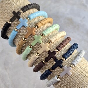 Op de afbeelding: Een collectie van tien elastische armbanden gemaakt van platte, ronde kleikralen in diverse kleuren zoals zwart, lichtblauw, beige, mintgroen, cr&egrave;me, bruin, marineblauw en grijs, elk voorzien van een centraal kruisbedeltje en gouden kraaltjes.