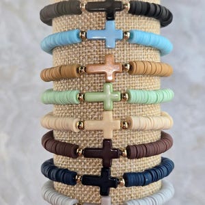 Op de afbeelding: Een stapel van negen elastische armbanden gemaakt van platte, ronde polymeerklei kralen in diverse kleuren zoals zwart, lichtblauw, beige, mintgroen, cr&egrave;me, bruin, marineblauw en grijs, elk met een centraal kruisbedeltje en goudkleurige kralen.
