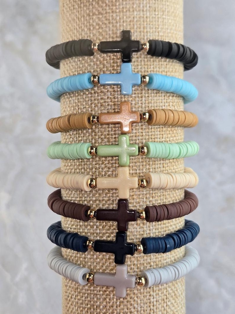 Op de afbeelding: Een stapel van negen elastische armbanden gemaakt van platte, ronde polymeerklei kralen in diverse kleuren zoals zwart, lichtblauw, beige, mintgroen, cr&egrave;me, bruin, marineblauw en grijs, elk met een centraal kruisbedeltje en goudkleurige kralen.