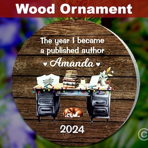 Op de afbeelding: Een ronde houten ornament met een bruine houtnerf achtergrond. De ornament heeft een bureau met een laptop, boeken en een hond. De tekst op de ornament luidt "The year I became a published author Amanda 2024".