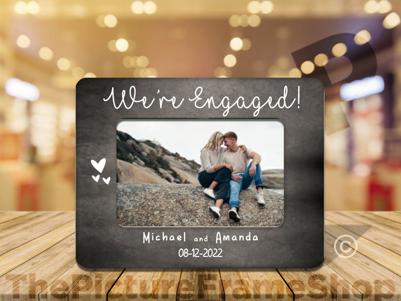 May include: A black chalkboard photo frame with a white inscription "We're Engaged!" and a photo of a couple. The frame also includes the names "Michael and Amanda" and the date "08-12-2022".