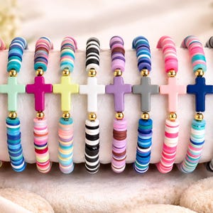 May include: A collection of colorful beaded bracelets with cross pendants. Each bracelet features a different color scheme, including red, blue, pink, and black, with gold-colored accents. The bracelets are displayed on a white surface.