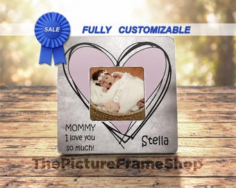 May include: A square photo frame with a light gray background featuring a large pink heart outline and black scribbled heart design. The frame displays a photo of a baby in a basket, with the text "MOMMY I love you so much!" and "Stella".