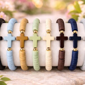 Op de afbeelding: Acht elastische armbanden op een rij, elk voorzien van platte ronde kleikralen in diverse effen kleuren waaronder zwart, lichtblauw, beige, lichtgroen, cr&egrave;me, bruin, marineblauw en grijs, in het midden een bijpassend kruis en gouden kralen.