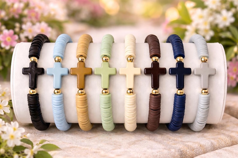 Op de afbeelding: Acht elastische armbanden op een rij, elk voorzien van platte ronde kleikralen in diverse effen kleuren waaronder zwart, lichtblauw, beige, lichtgroen, cr&egrave;me, bruin, marineblauw en grijs, in het midden een bijpassend kruis en gouden kralen.