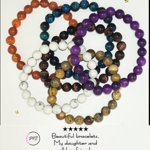 Op de afbeelding: Een collectie van vijf ronde kralenarmbanden met gladde, ronde stenen in diverse kleuren, waaronder oranje, blauw, paars, wit en bruin. Onder de armbanden staat de tekst: 5 sterren, Beautiful bracelets. My daughter and all her friends love them!