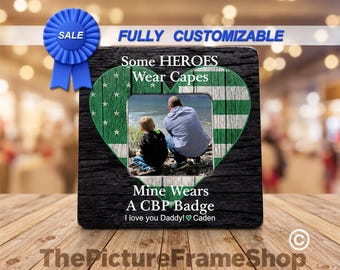 May include: A square photo frame with a black wood grain texture featuring a green heart graphic with white stars and stripes. The text reads: Some HEROES Wear Capes, Mine Wears A CBP Badge, I love you Daddy! Caden. A blue ribbon icon is at the top left.
