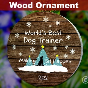 May include: Round wooden Christmas ornament with a brown woodgrain background. The ornament features a white silhouette of a dog sitting in front of two blue Christmas trees with yellow stars on top. The text "World's Best Dog Trainer" is written in white above the trees. The text "Making Sit Happen" is written in white below the trees. The year "2022" is written in white at the bottom of the ornament.