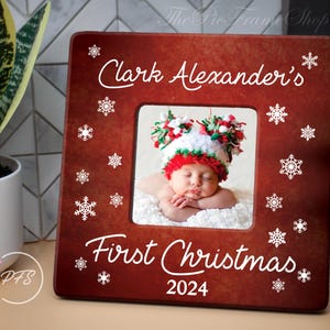 Peut inclure: Cadre photo en bois rouge avec des flocons de neige blancs et le texte "Clark Alexander's First Christmas 2024". Une photo d'un b&eacute;b&eacute; portant un chapeau rouge et blanc est dans le cadre.