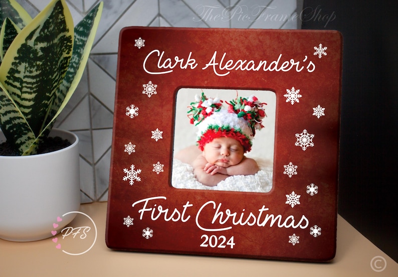 Peut inclure: Cadre photo en bois rouge avec des flocons de neige blancs et le texte "Clark Alexander's First Christmas 2024". Une photo d'un b&eacute;b&eacute; portant un chapeau rouge et blanc est dans le cadre.