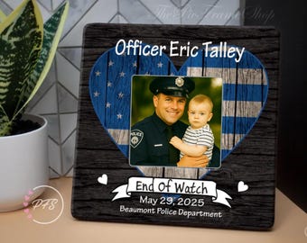 Op de afbeelding: Een vierkante herdenkingsplaat met een donkere houtnerfachtergrond met een blauw hart met een Thin Blue Line vlagpatroon. Het toont een foto van een agent die een baby vasthoudt, met de tekst Officer Eric Talley, End Of Watch, May 29, 2025, en Beaumont Police Department.