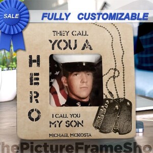 Op de afbeelding: Een vierkante houten fotolijst met een lichtbruine afwerking. De lijst bevat de tekst "THEY CALL YOU A HERO I CALL YOU MY SON" en "MICHAEL MCKOSTA" met een afbeelding van twee militaire dogtags. Een blauw verkooplint is bevestigd.