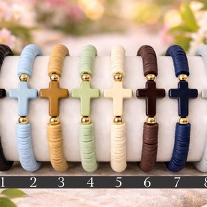 Op de afbeelding: Acht elastische armbanden op een rij, elk voorzien van platte ronde kleikralen in diverse kleuren zoals zwart, blauw, beige, groen, cr&egrave;me, bruin, donkerblauw en grijs, met een centrale kruiskraal geflankeerd door twee kleine goudkleurige kralen.
