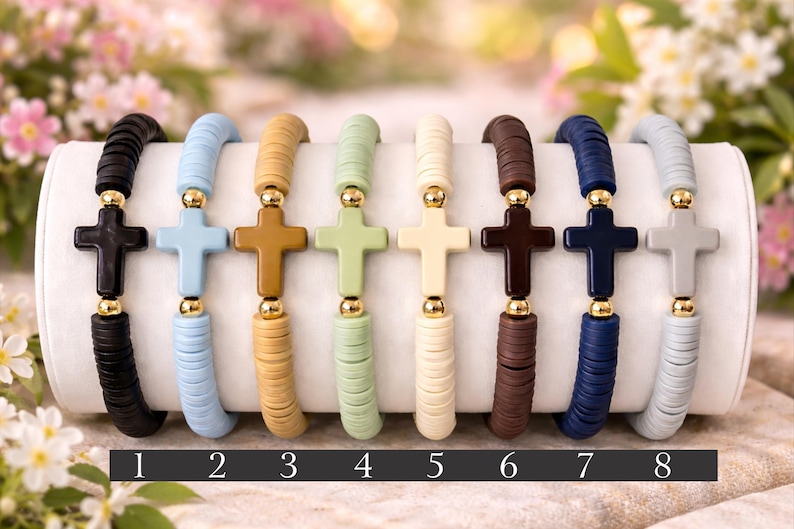 Op de afbeelding: Acht elastische armbanden op een rij, elk voorzien van platte ronde kleikralen in diverse kleuren zoals zwart, blauw, beige, groen, cr&egrave;me, bruin, donkerblauw en grijs, met een centrale kruiskraal geflankeerd door twee kleine goudkleurige kralen.