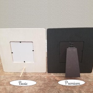 May include: Two picture frames, one white and one black, both with a simple design and a small opening for a photo. The white frame has a wooden stand, while the black frame has a more substantial stand.