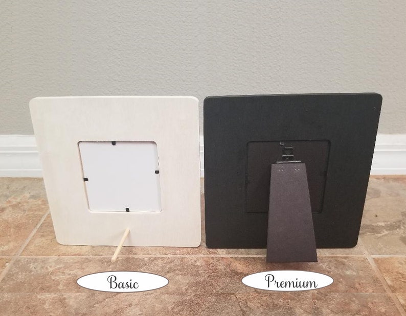 May include: Two picture frames, one white and one black, both with a simple design and a small opening for a photo. The white frame has a wooden stand, while the black frame has a more substantial stand.