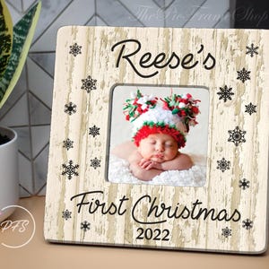 Peut inclure: Un cadre photo en bois blanc avec une finition vieillie. Le cadre est d&eacute;cor&eacute; d'un motif de flocons de neige noirs et du texte "Reese's First Christmas 2022".