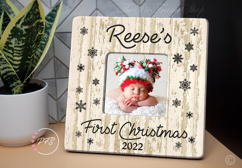 Peut inclure: Un cadre photo en bois blanc avec une finition vieillie. Le cadre est d&eacute;cor&eacute; d'un motif de flocons de neige noirs et du texte "Reese's First Christmas 2022".